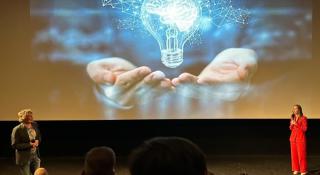 Twee mensen op een podium, met een projectie van een lamp op de achtergrond